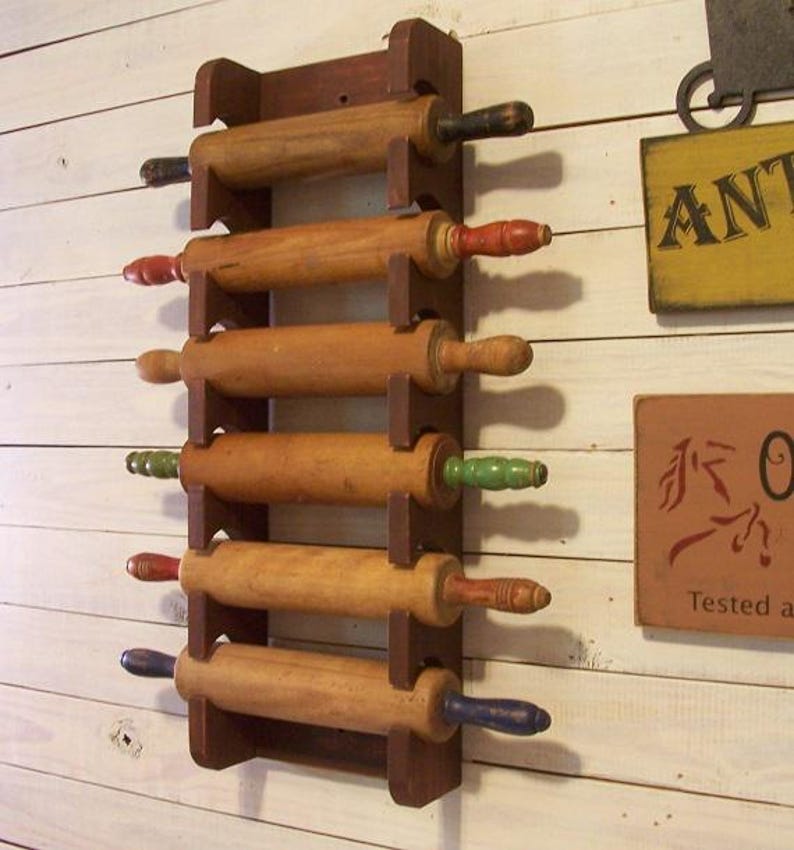 Op de afbeelding: Een houten deegrollerrek met zes deegrollers. De deegrollers hebben gekleurde handvatten in rood, groen, blauw en zwart. Het rek is donkerbruin en is gemonteerd op een witte houten muur.