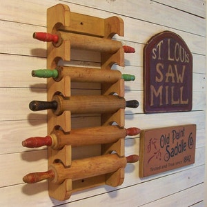 Peut inclure: Un porte-rouleaux en bois avec cinq rouleaux &agrave; p&acirc;tisserie. Le support est peint en jaune et a des poign&eacute;es rouges et vertes. Le support est mont&eacute; sur un mur en bois blanc. Il y a deux panneaux sur le mur, l'un qui dit "St. Louis Saw Mill" et l'autre qui dit "Old Paint Saddle Co. Tested and True since 1842".