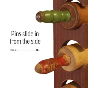 Op de afbeelding: Een houten deegrollerhouder met drie deegrollers. De deegrollers hebben groene, rode en natuurlijke houten handvatten. De houder is donkerbruin met cirkelvormige openingen. De tekst aan de linkerkant luidt "Pins slide in from the side".