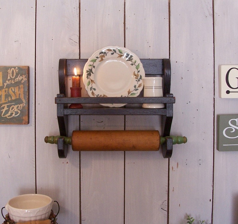 May include: A dark brown wooden wall shelf featuring a top tier holding a small lit candle, a decorative plate with a leaf pattern, and a white mug. Below the shelf, a wooden rolling pin with green handles is suspended on two brackets.