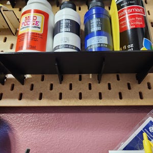 Może przedstawiać: Czarna półka z czterema butelkami farby i puszką ze sprayem. Butelki są oznaczone "Mod Podge", "Liquitex Basics Acrylic Fluid" i "Liquitex Basics Acrylic Fluid". Puszka ze sprayem jest oznaczona "Craft Smart Matte Acrylic Paint".
