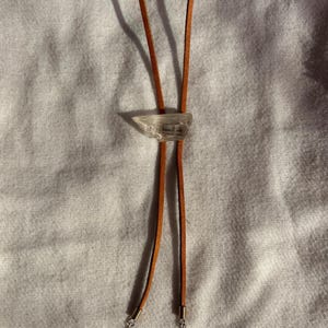 May include: A bolo tie featuring a clear, raw-cut crystal centerpiece mounted on a tan leather cord. The two ends of the cord are finished with silver-toned metal coils and small, pale yellow beaded accents, creating a simple, functional accessory.