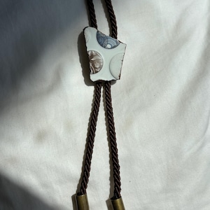 May include: A bolo tie featuring a central piece of white ceramic with circular blue and brown floral patterns. The cord is made of dark brown twisted rope, and the two ends are finished with metallic brass-colored bullet casing tips.