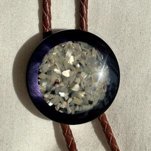 May include: A bolo tie featuring a circular pendant with a dark purple metallic rim and a center inlay of small, iridescent, off-white crushed shell fragments. The pendant is attached to a braided brown leather cord that extends upward and downward.
