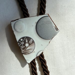 May include: A bolo tie featuring a white ceramic shard pendant with a brown and gray circular pattern. The pendant displays a large gray circle, a smaller white circle, and a circle with intricate brown floral line art. It hangs from a dark brown cord.