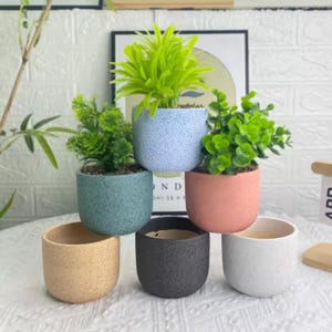 May include: A collection of small, colorful ceramic pots, some filled with artificial greenery. The pots are in shades of blue, green, peach, beige, black, and white. The pots have a speckled texture and are arranged in a stacked display.