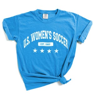 May include: A light blue t-shirt with white text that reads "U.S. WOMEN'S SOCCER EST. 1985" with three white stars below the text.