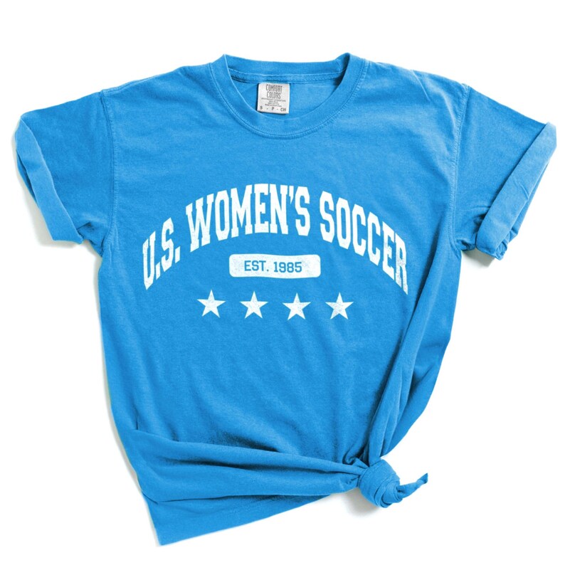 May include: A light blue t-shirt with white text that reads "U.S. WOMEN'S SOCCER EST. 1985" with three white stars below the text.