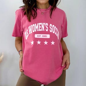 May include: Pink t-shirt with a white graphic that says "US Women's Soccer Est. 1985" with three white stars underneath.