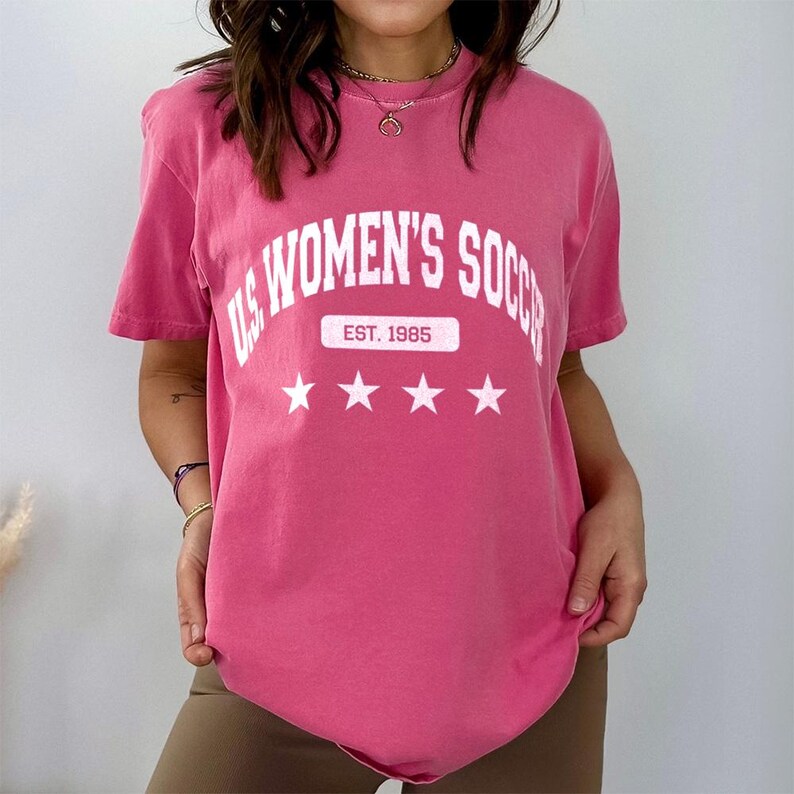 May include: Pink t-shirt with a white graphic that says "US Women's Soccer Est. 1985" with three white stars underneath.