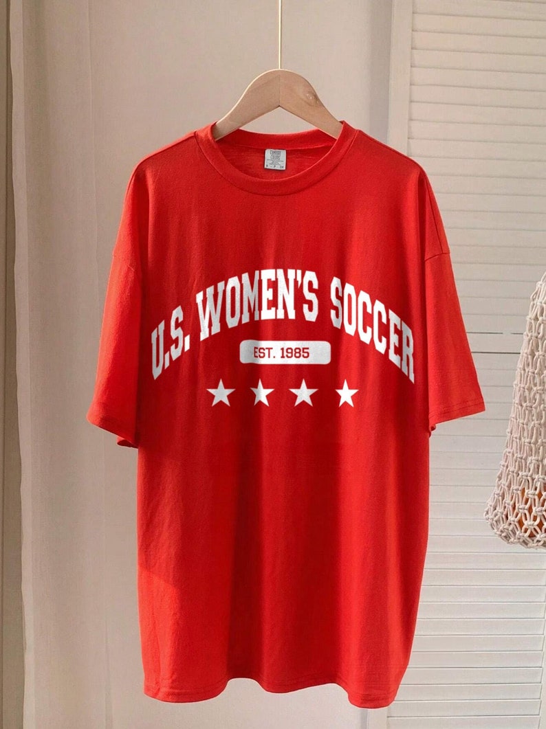 May include: Red t-shirt with white lettering that says "U.S. Women's Soccer" with a banner that says "Est. 1985" and four white stars.