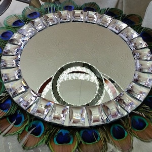 May include: A round mirror with a decorative border of faux peacock feathers and clear rhinestones. The mirror is surrounded by a ring of iridescent blue and green feathers.