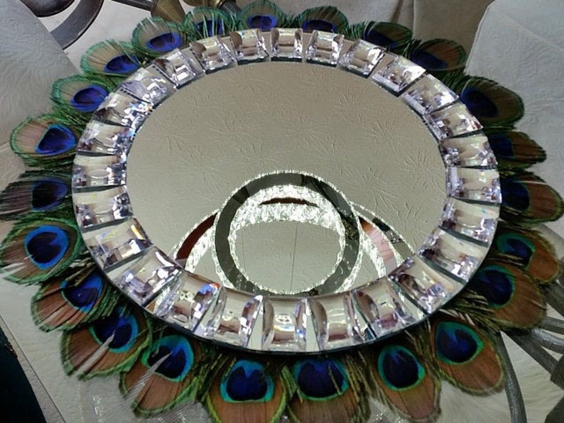 May include: A round mirror with a decorative border of faux peacock feathers and clear rhinestones. The mirror is surrounded by a ring of iridescent blue and green feathers.