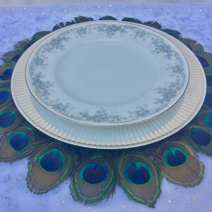 May include: A white dinner plate with a blue floral pattern sits on top of a silver charger plate. The plates are centered on a peacock feather charger, which is a circular arrangement of peacock feathers with green, blue, and brown colors.