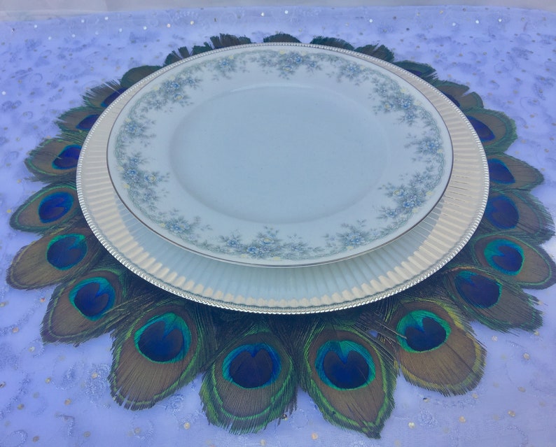 May include: A white dinner plate with a blue floral pattern sits on top of a silver charger plate. The plates are centered on a peacock feather charger, which is a circular arrangement of peacock feathers with green, blue, and brown colors.