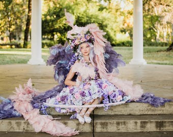May include: A person wearing a fantasy costume with a large feathered headdress and skirt. The costume features shades of purple, pink, and blue feathers and flowers. The person is sitting on steps in front of a white columned structure.