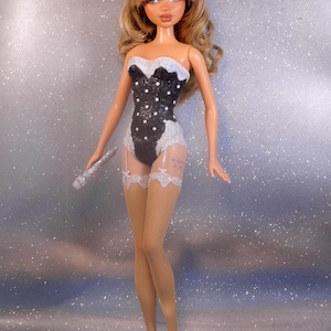 May include: A fashion doll with long wavy blonde hair, wearing a black strapless bodysuit with white polka dots and white lace trim. The doll features white garter straps, sheer tan fishnet stockings, and white heels, while holding a silver microphone.