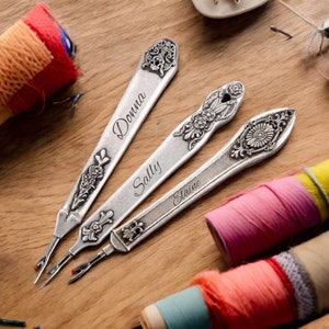 May include: Three silver seam rippers with ornate floral designs and personalized names: "Donna", "Sally", and "Elaine". The rippers are on a wooden surface, with spools of colorful thread in the background. A sewing-themed item.
