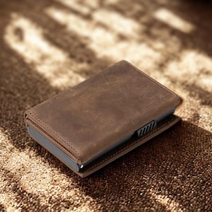 May include: A brown leather wallet with a textured surface. The wallet is rectangular and has a card holder mechanism on the side. The wallet is sitting on a textured brown surface.