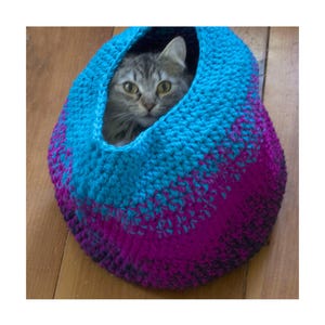 May include: A grey tabby cat is nestled inside a handmade crochet cat bed. The bed is crafted from blue and purple yarn, providing a cosy and colourful space for a pet. The cat's eyes are fixed forward.