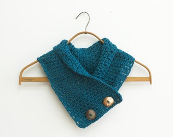 May include: Teal blue crocheted cowl with two brown buttons. The cowl is hanging on a wooden hanger.