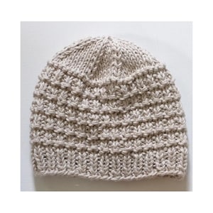 May include: A beige knit beanie featuring a thick ribbed brim at the base and a textured, repeating stitch pattern across the main body. The hat is constructed from chunky yarn, showcasing a rounded crown and a consistent, neutral light tan color.