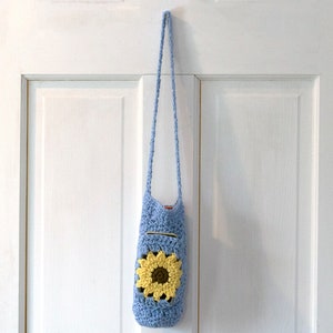 May include: A light blue crocheted bag with a sunflower design. The bag has a long strap and is hanging on a white door.
