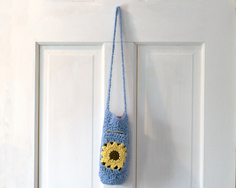 May include: A light blue crocheted bag with a sunflower design. The bag has a long strap and is hanging on a white door.