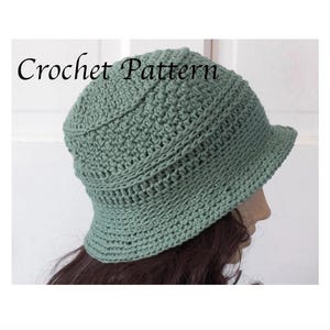 May include: A sage green bucket hat made with a textured crochet stitch pattern. The hat features a rounded crown and a downward-sloping brim. The text "Crochet Pattern" is printed in black letters across the upper portion of the image.