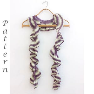 Peut inclure: Une écharpe en crochet rayée de violet et de blanc avec des bords festonnés. L'écharpe est suspendue à un cintre en bois.