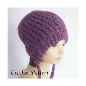 Könnte beinhalten: Eine gehäkelte lila Mütze mit Ohrenklappen und geflochtenen Bändern. Die Mütze ist mit einem strukturierten Stich gefertigt und trägt die Worte "Crochet Pattern" in der unteren linken Ecke.