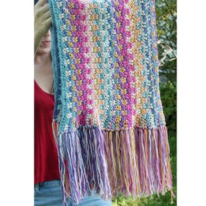 May include: A colorful, handmade crochet scarf with a fringed edge. The scarf features a mosaic pattern in shades of blue, pink, yellow, and white. The fringe is a mix of purple, white, and yellow. This scarf is a cozy accessory.