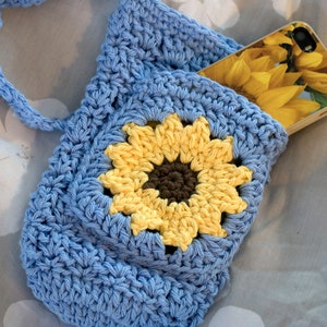 May include: A blue crocheted bag with a sunflower design. The bag has a long strap and a pocket on the front. A phone with a sunflower case is in the pocket.
