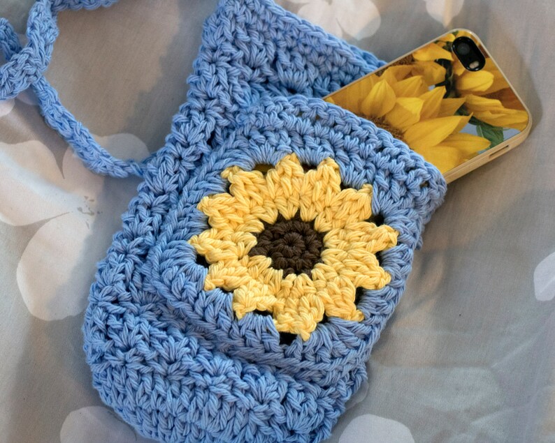 May include: A blue crocheted bag with a sunflower design. The bag has a long strap and a pocket on the front. A phone with a sunflower case is in the pocket.