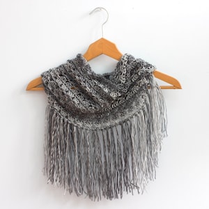Peut inclure: Une écharpe en crochet grise avec des franges qui pendent du bord inférieur. L'écharpe est présentée sur un cintre en bois sur un fond blanc.