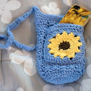 May include: A blue crocheted phone pouch with a sunflower design and a long strap. The pouch is holding a phone with a sunflower design case.
