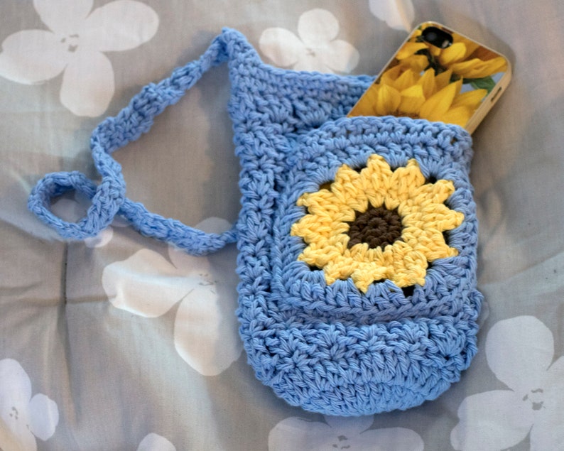 May include: A blue crocheted phone pouch with a sunflower design and a long strap. The pouch is holding a phone with a sunflower design case.