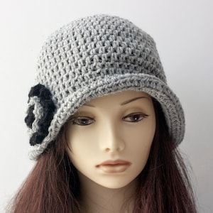 May include: A gray crocheted cloche hat with a black flower accent. The hat has a wide brim and a fitted crown.