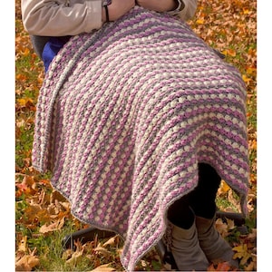 May include: A handmade crochet blanket in a square pattern. The blanket features alternating rows of gray, cream, and pink. The blanket is draped over a person's lap, with fall leaves in the background.