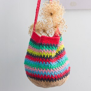 May include: A colorful crocheted tote bag with a striped pattern in red, green, blue, pink, and yellow. The bag is hanging from a white wall with dried flowers inside.