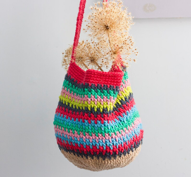 May include: A colorful crocheted tote bag with a striped pattern in red, green, blue, pink, and yellow. The bag is hanging from a white wall with dried flowers inside.