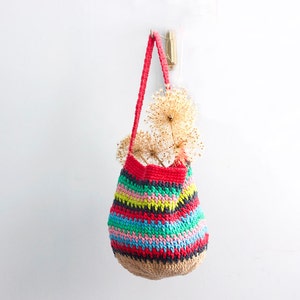 May include: A colorful striped crocheted tote bag with a natural jute bottom, hanging from a hook on a white wall. The bag is filled with dried flowers.
