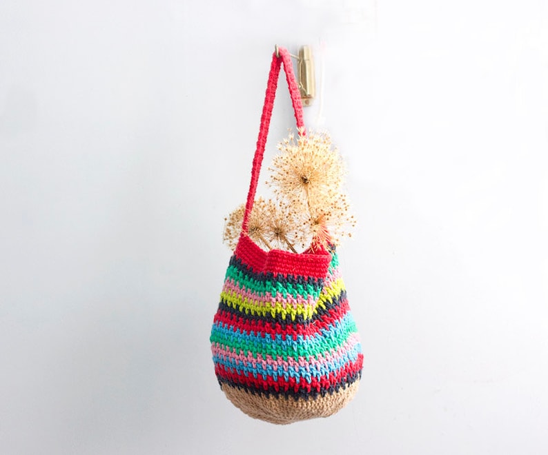 May include: A colorful striped crocheted tote bag with a natural jute bottom, hanging from a hook on a white wall. The bag is filled with dried flowers.