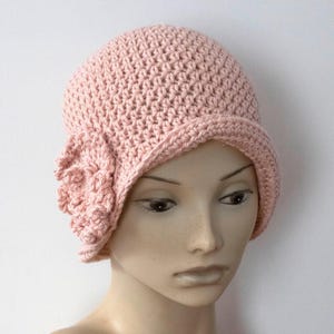 May include: A light pink crochet cloche hat with a small brim and a decorative flower on the side. The hat is made of a soft, textured yarn and is a stylish accessory.