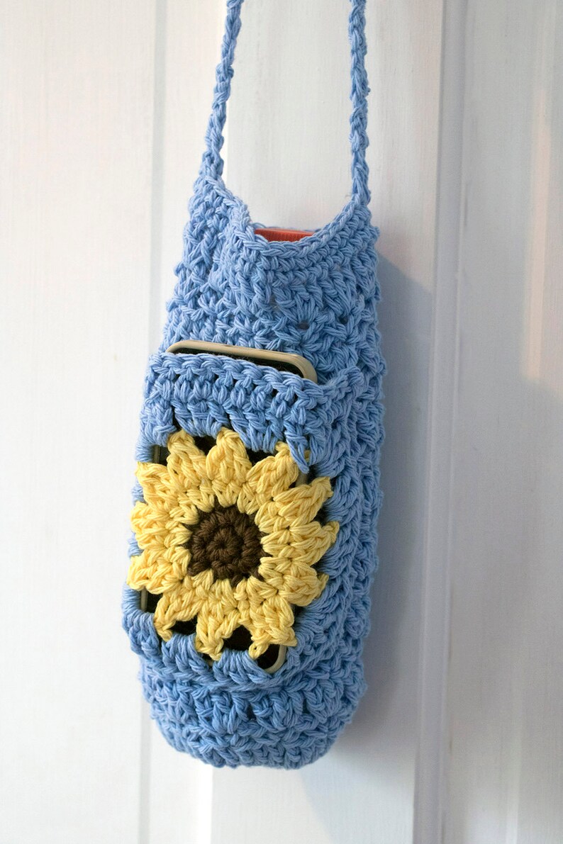May include: A light blue crocheted bag with a sunflower design. The bag has a pocket on the front and a long strap.