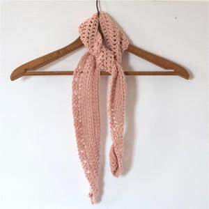 May include: A light pink, crocheted scarf hanging on a wooden hanger. The scarf is made of a soft, open-weave material, with a long, slender design. The scarf is a solid color and appears to be handmade.