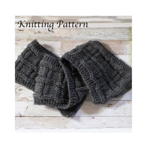 May include: A dark grey knitted scarf with a textured pattern, displayed on a light wood surface. The scarf is folded and draped, showcasing its woven design. The words "Knitting Pattern" are written at the top.