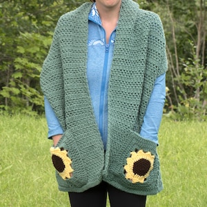 May include: A green crochet shawl with two pockets featuring crocheted sunflower designs. The shawl is worn over a blue shirt.