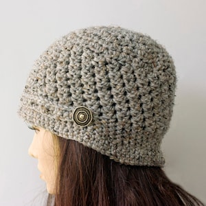 May include: A gray crocheted beanie with a brown button on the side. The beanie is worn on a mannequin with long brown hair.