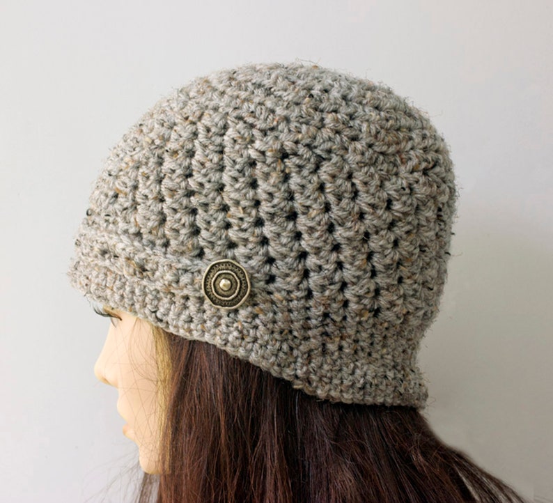 May include: A gray crocheted beanie with a brown button on the side. The beanie is worn on a mannequin with long brown hair.